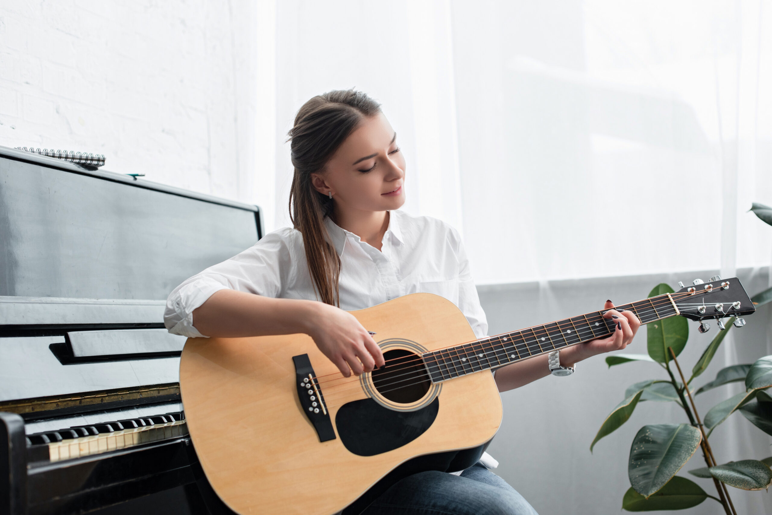 attractive girl sitting and playing guitar in living room with piano on background