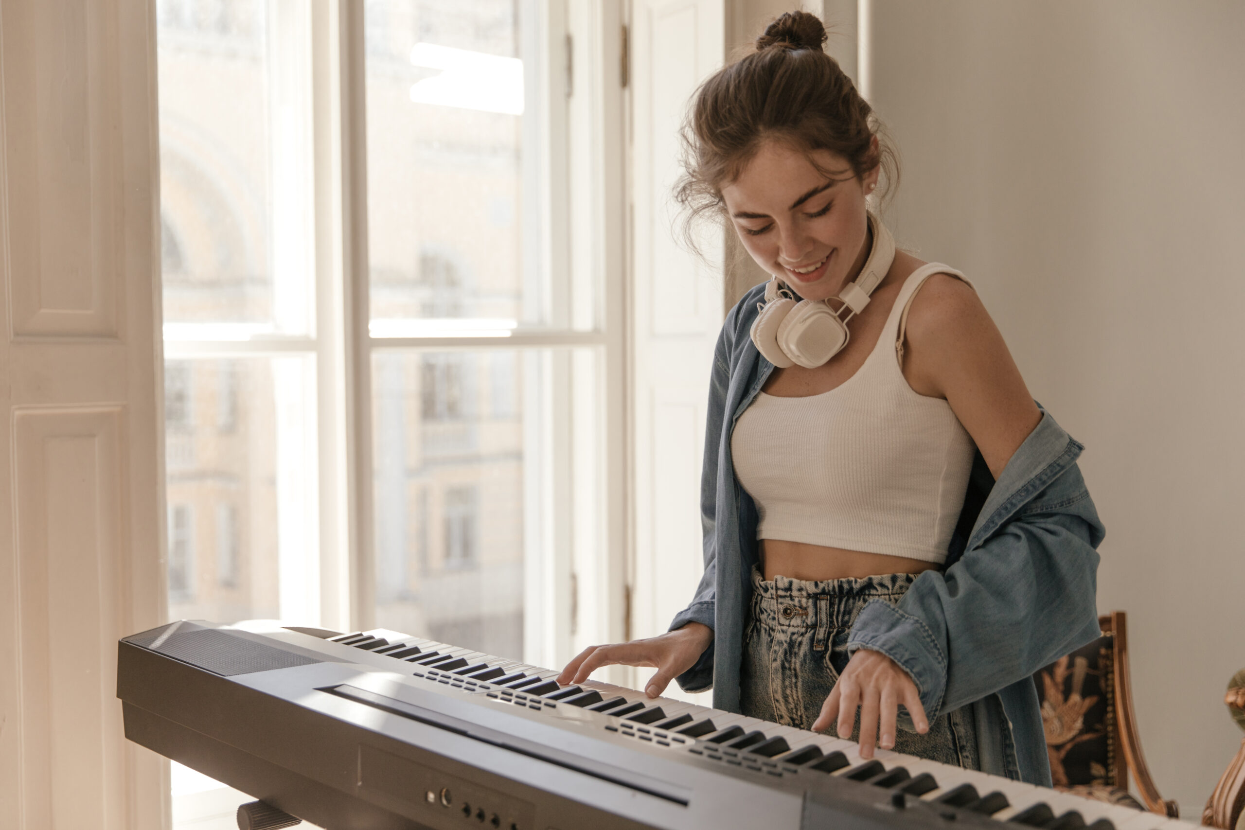 Cute young student playing piano at home near window. Attractive girl with collected hair, white to